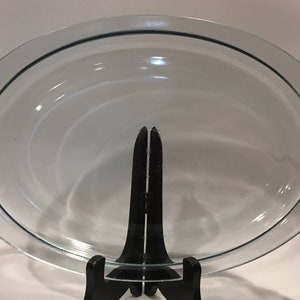 Könnte beinhalten: Klarer, ovaler Glasbackform mit gewelltem Rand und Griffen. Die Form wird von einem schwarzen Ständer getragen. Die Form ist etwa 30 cm lang und 20 cm breit.