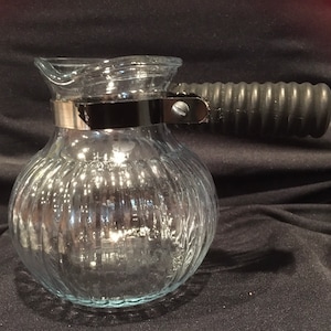 May include: Clear glass coffee pot with a ribbed design. It features a black handle and a silver metal band around the neck. The pot is designed for brewing coffee and has a spout for easy pouring.