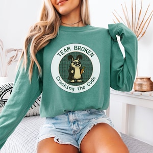 May include: Teal green sweatshirt with a circular graphic. The graphic features a cartoon rabbit wearing sunglasses and holding a carrot, with the text "TEAM BROKEN Cracking the Code" around the edge. The rabbit is set against a background of binary code.