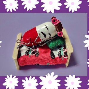 Op de afbeelding: Een rood en wit pilvormig figuurtje met een geschilderd gezicht en bruin haar ligt op een miniatuurbed met een rood-wit gepatroneerd dekbed en een groen kussen.