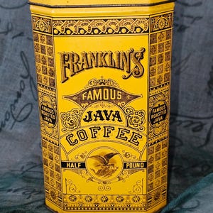 Könnte beinhalten: Eine gelbe, sechseckige Metalldose mit schwarzen Zierrändern und der Aufschrift FRANKLIN'S FAMOUS JAVA COFFEE. Die Vorderseite zeigt eine Adlerillustration, die Worte HALF POUND und zwei runde Abzeichen mit der Aufschrift FAMOUS ROASTED JAVA.