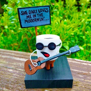 Op de afbeelding: Een witte koffiemok met een zwarte snor en zonnebril houdt een bruine gitaar vast en een bord met de tekst "Ze houdt alleen van me in de ochtend...".