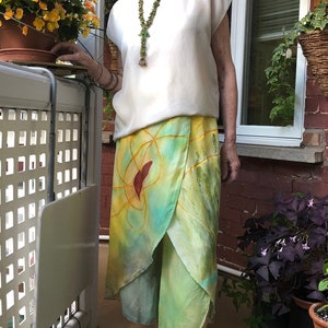 Könnte beinhalten: Eine Frau trägt ein weißes Oberteil mit einer grünen Perlenkette und einen grün-gelb-blauen Wickelrock mit rotem Muster. Sie steht auf einer Terrasse mit Pflanzen im Hintergrund.