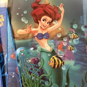 Puede incluir: Una bolsa de regalo rectangular con una colorida escena submarina que muestra a una sirena de pelo rojo con un top de conchas moradas y una cola verde, rodeada por una medusa azul, peces de rayas amarillas y negras y burbujas flotantes.