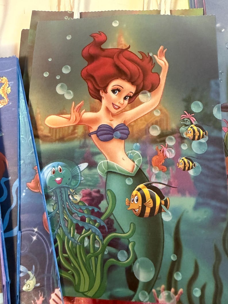 Puede incluir: Una bolsa de regalo rectangular con una colorida escena submarina que muestra a una sirena de pelo rojo con un top de conchas moradas y una cola verde, rodeada por una medusa azul, peces de rayas amarillas y negras y burbujas flotantes.