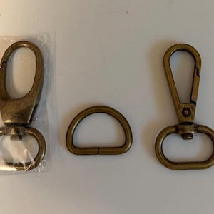 May include: Two antique brass-toned metal swivel lobster claw clasps and one matching D-ring. One clasp is enclosed in a clear plastic bag, while the other clasp and the D-ring are positioned outside of any packaging on a plain surface.
