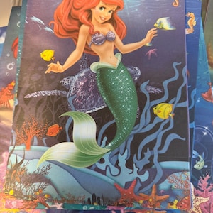 Puede incluir: Una bolsa de regalo rectangular de papel con una colorida escena submarina que muestra a una sirena con cabello largo y rojo, un top de conchas moradas y una cola verde brillante. El fondo representa un oc&eacute;ano azul profundo con una tortuga marina, varios peces tropicales, arrecifes de coral y estrellas de mar.