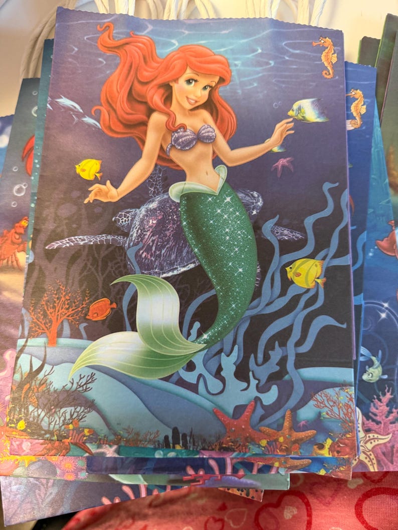 Puede incluir: Una bolsa de regalo rectangular de papel con una colorida escena submarina que muestra a una sirena con cabello largo y rojo, un top de conchas moradas y una cola verde brillante. El fondo representa un oc&eacute;ano azul profundo con una tortuga marina, varios peces tropicales, arrecifes de coral y estrellas de mar.
