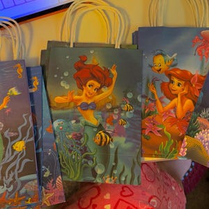 Puede incluir: Una colecci&oacute;n de bolsas de regalo de papel con ilustraciones de la sirena Ariel de La Sirenita. Cada bolsa muestra una escena submarina diferente con corales, vida marina y burbujas en tonos azules, verdes y rojos.