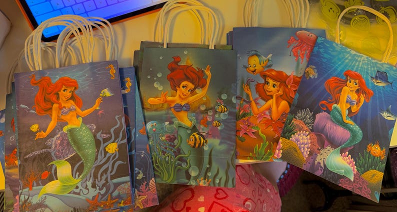 Puede incluir: Una colecci&oacute;n de bolsas de regalo de papel con ilustraciones de la sirena Ariel de La Sirenita. Cada bolsa muestra una escena submarina diferente con corales, vida marina y burbujas en tonos azules, verdes y rojos.