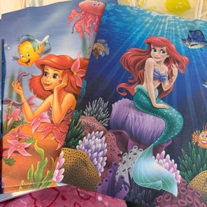 Puede incluir: Dos bolsas de regalo de papel con personajes de La Sirenita. Una bolsa muestra a Ariel con Flounder y una medusa sobre un fondo azul claro. La otra bolsa representa a Ariel con cabello rojo y cola verde sentada en un arrecife de coral morado.
