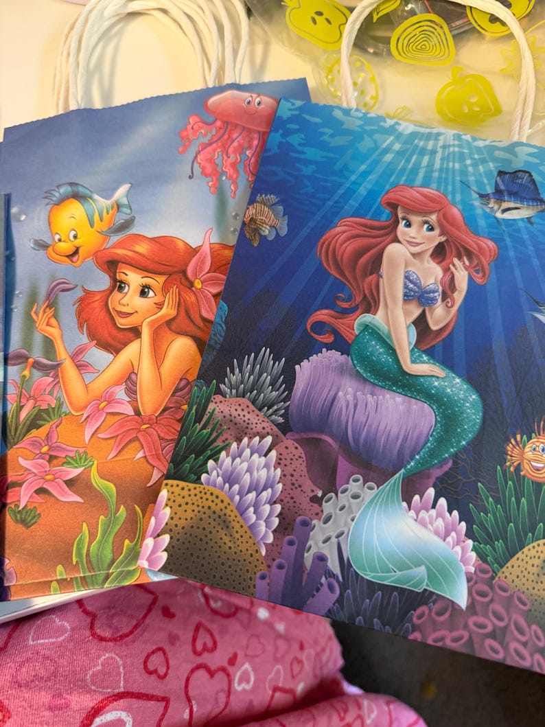 Puede incluir: Dos bolsas de regalo de papel con personajes de La Sirenita. Una bolsa muestra a Ariel con Flounder y una medusa sobre un fondo azul claro. La otra bolsa representa a Ariel con cabello rojo y cola verde sentada en un arrecife de coral morado.