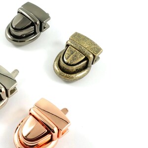 May include: Close-up of four metal purse clasps in various finishes: gunmetal, antique brass, silver, and rose gold. The clasps are oval-shaped with a rectangular top, designed for securing bags or other accessories. The background is white.