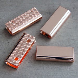 May include: Four rectangular rose gold metal cord ends. Two of the pieces feature a textured grid pattern with two small circular holes, while the other two pieces have a smooth, polished finish. Each piece is folded into a U-shape channel.