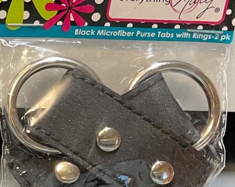 May include: Black microfiber purse tabs with silver rings and snap closures. The package includes two tabs, as indicated by the text on the label. The tabs are designed to attach to a purse or bag.