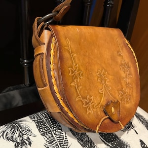 May include: A brown leather satchel bag with a floral design and a braided strap. The bag has a flap closure and a metal ring.
