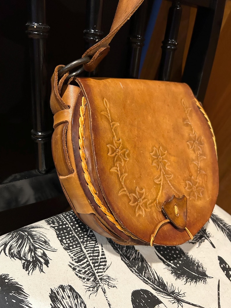 May include: A brown leather satchel bag with a floral design and a braided strap. The bag has a flap closure and a metal ring.