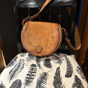 May include: A brown leather satchel bag with a floral design. The bag has a long strap and a flap closure. The bag is sitting on a chair with a white and black feather pattern.