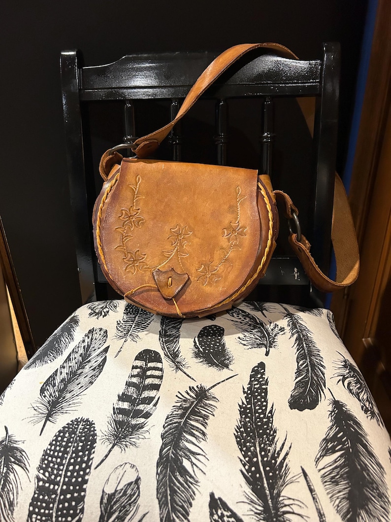 May include: A brown leather satchel bag with a floral design. The bag has a long strap and a flap closure. The bag is sitting on a chair with a white and black feather pattern.