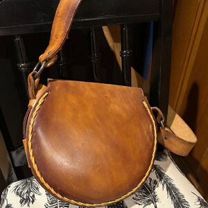 May include: A brown leather satchel bag with a circular flap and a long strap. The bag has a stitched detail around the edge of the flap.