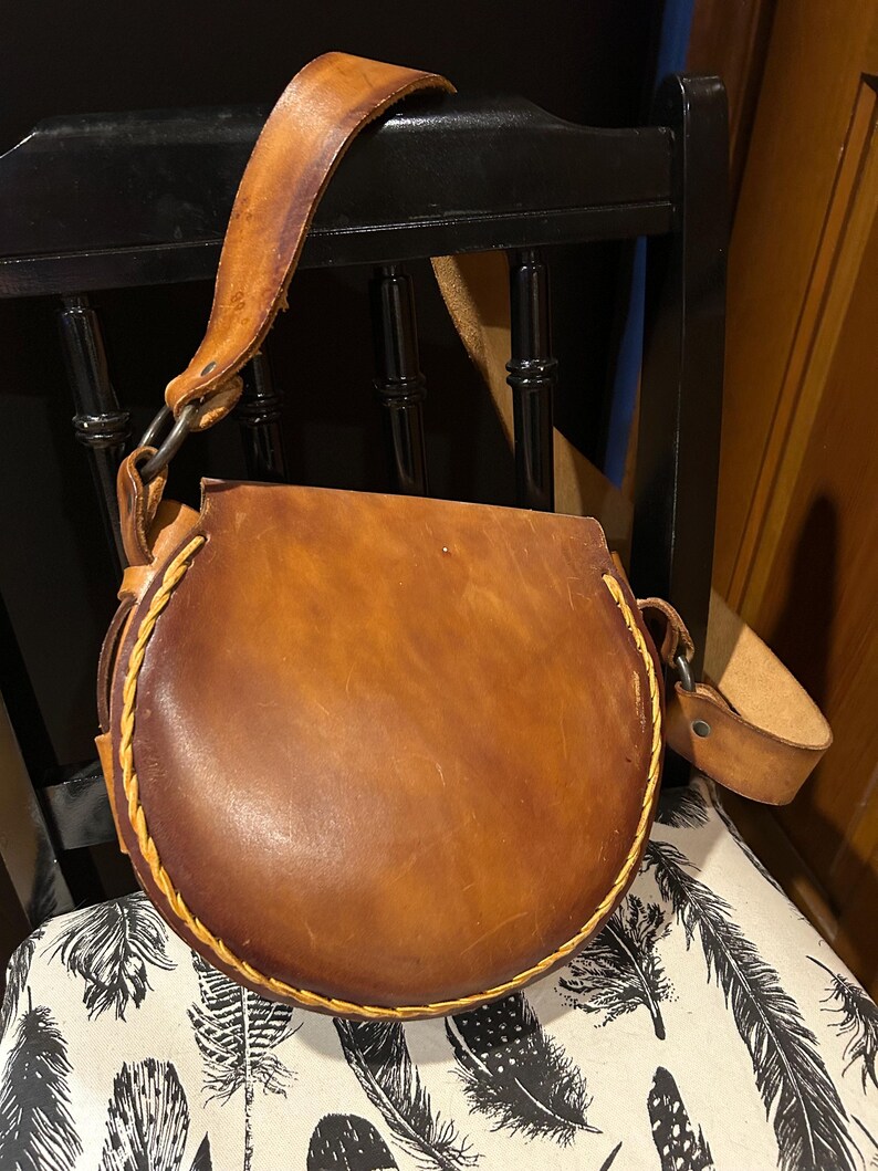 May include: A brown leather satchel bag with a circular flap and a long strap. The bag has a stitched detail around the edge of the flap.