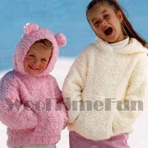 May include: Two children wearing knitted hooded sweatshirts. The girl on the left is wearing a pink hooded sweatshirt with bear ears. The girl on the right is wearing a white hooded sweatshirt. Both girls are wearing jeans.