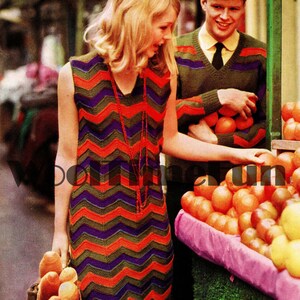 May include: A woman wearing a sleeveless dress with a chevron pattern in orange, green, and purple. She is holding a loaf of bread and smiling. A man wearing a green sweater with a chevron pattern is standing behind her.