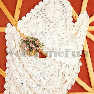 May include: A white crocheted baby blanket with a delicate floral pattern. The blanket is laid out on a wooden surface with a small bouquet of pink flowers.