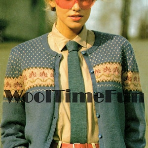 May include: A blue knitted cardigan with a white and pink patterned design. The cardigan has a button closure and a tie neck. The model is wearing a white shirt with a blue tie and beige trousers.