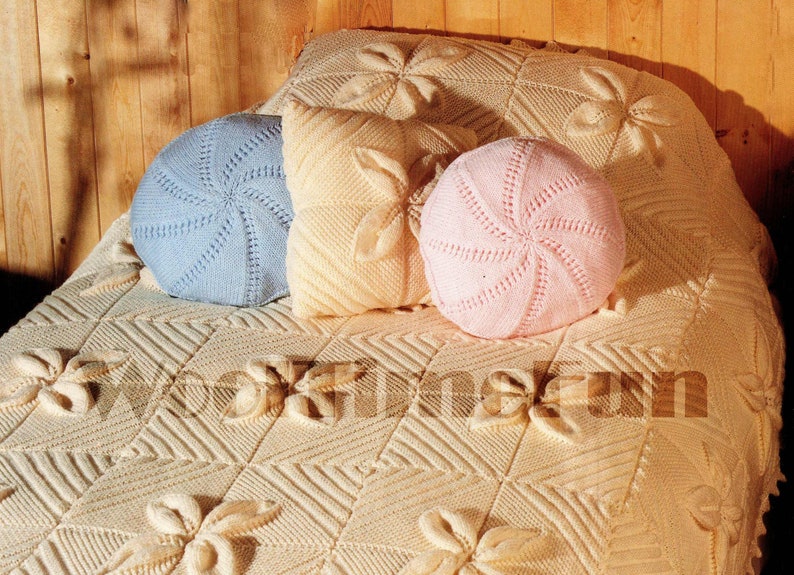 May include: A white knitted throw blanket with a floral pattern and three knitted pillows, one blue, one pink, and one cream.