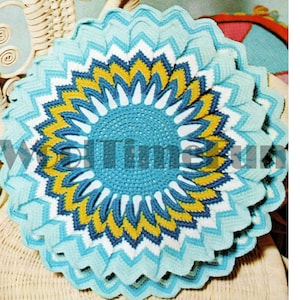 Puede incluir: Un cojín redondo de ganchillo en tonos azules, blancos y amarillos. El cojín presenta un diseño circular central con patrones radiales, creando una apariencia de flor. El borde exterior tiene un patrón en zigzag.