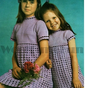 May include: Two girls wearing matching purple crochet dresses. The dresses have a lacy pattern and a scalloped hem. The girls are holding a bouquet of flowers.