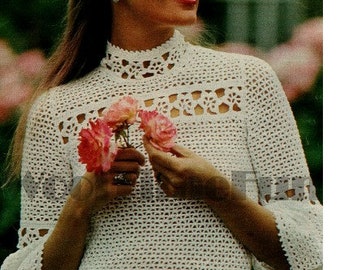 Pode incluir: Um top branco de crochê com gola alta e mangas bufantes. O top apresenta um delicado padrão de renda e é usado por uma mulher segurando flores cor-de-rosa.