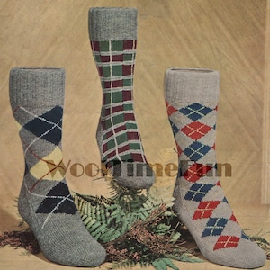 May include: Three pairs of grey socks with different colored patterns. The socks on the left have a diamond pattern with red, yellow, and blue accents. The socks in the middle have a plaid pattern with red, green, and white accents. The socks on the right have a diamond pattern with red, blue, and white accents. The socks are sitting on a brown surface with green ferns.