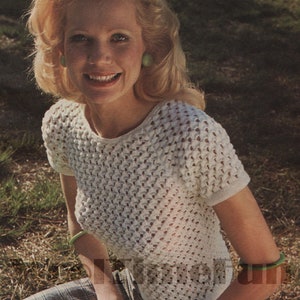 May include: A woman with blonde hair is wearing a white crocheted top with a delicate openwork pattern. She is sitting on a plaid skirt and has green earrings and bracelets.