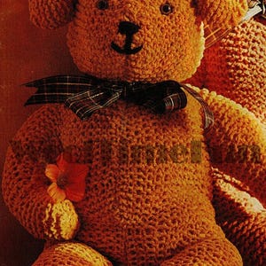 Op de afbeelding: Een gehaakte teddybeer met een geruite strik en een bloem in zijn poot. De beer is bruin en heeft zwarte ogen en een zwarte neus.