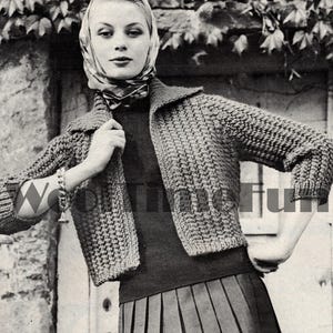 Può includere: Una foto in bianco e nero di una donna che indossa un cardigan all'uncinetto con una sciarpa legata intorno alla testa. Il cardigan ha un colletto e maniche lunghe. La donna è in piedi davanti a un muro di pietra con l'edera che cresce su di esso.