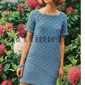 May include: A woman wearing a light blue crochet dress with a scalloped neckline. She is standing in front of a background of pink and green flowers.