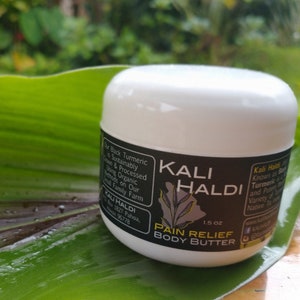 Puede incluir: Un tarro blanco de Kali Haldi Pain Relief Body Butter, 42,5 g, descansa sobre una gran hoja verde. El tarro tiene una etiqueta negra con texto blanco y una ilustración de una planta. El fondo es follaje verde borroso.