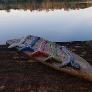 Puede incluir: Cinco adornos de cola de sirena de cristal de colores azul, rojo y verde, con detalles de purpurina. Están colocados sobre una paleta de madera, con un lago sereno de fondo.