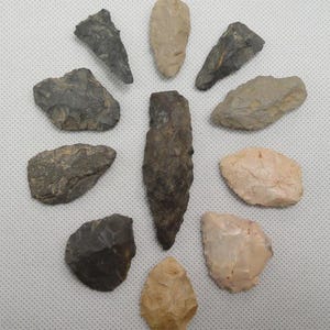 May include: A collection of various hand-chipped stone tools, including arrowheads and blades, in shades of gray, brown, and beige. The tools are arranged in a circular pattern, showcasing their different shapes and sizes. These primitive tools are likely from an archaeological find.