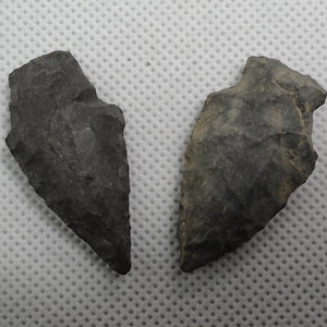 May include: Two dark gray stone arrowheads with chipped, serrated edges and pointed tips. Each arrowhead features a distinct triangular shape with a notched base, showcasing a rough, textured surface characteristic of knapped stone tools.