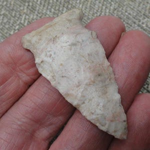 May include: A triangular stone arrowhead with a textured, off-white and light tan surface. The object features a pointed tip, serrated edges, and a notched base, displaying a natural, weathered appearance with visible mineral variations throughout.