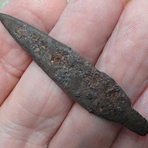 May include: A small, elongated, dark brown metal arrowhead with a heavily oxidized, pitted, and textured surface. The object features a sharp, pointed tip at one end and a flat, narrow tang at the base, showing significant signs of age and wear.