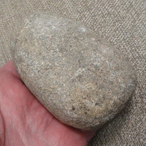 May include: A smooth, oval-shaped gray stone with a speckled surface. The stone is held in a hand, showcasing its natural texture and color variations. The stone's size is approximately 6 inches long.