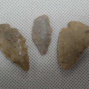 May include: Three ancient arrowheads in varying shades of tan and gray are displayed. The arrowheads have different shapes and sizes, showcasing the craftsmanship of early toolmakers. The textures range from smooth to slightly rough, highlighting the natural materials.