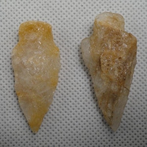 May include: Two arrowheads crafted from translucent, pale yellow and white quartz. Each piece features a pointed tip, sharp edges, and a notched base, displaying a crystalline, textured surface with natural mineral variations throughout.