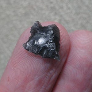 May include: A small, dark gray obsidian flake with a rough, fractured surface. The obsidian appears to be a natural stone, possibly a tool or artifact. The flake is held between two fingers, showing its size and texture.