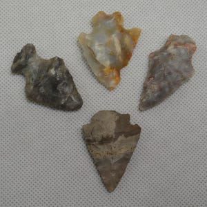 May include: Four ancient arrowheads in shades of gray, tan, and white are displayed. The arrowheads vary in shape and size, with visible textures and patterns. The background is a light gray, textured surface.