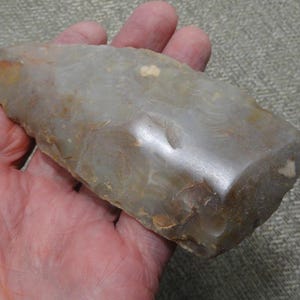 May include: A hand-knapped stone tool with a wedge-shaped profile, featuring a smooth, polished cutting edge and a textured, chipped surface in shades of light gray, tan, and brown, exhibiting natural mineral variations throughout.